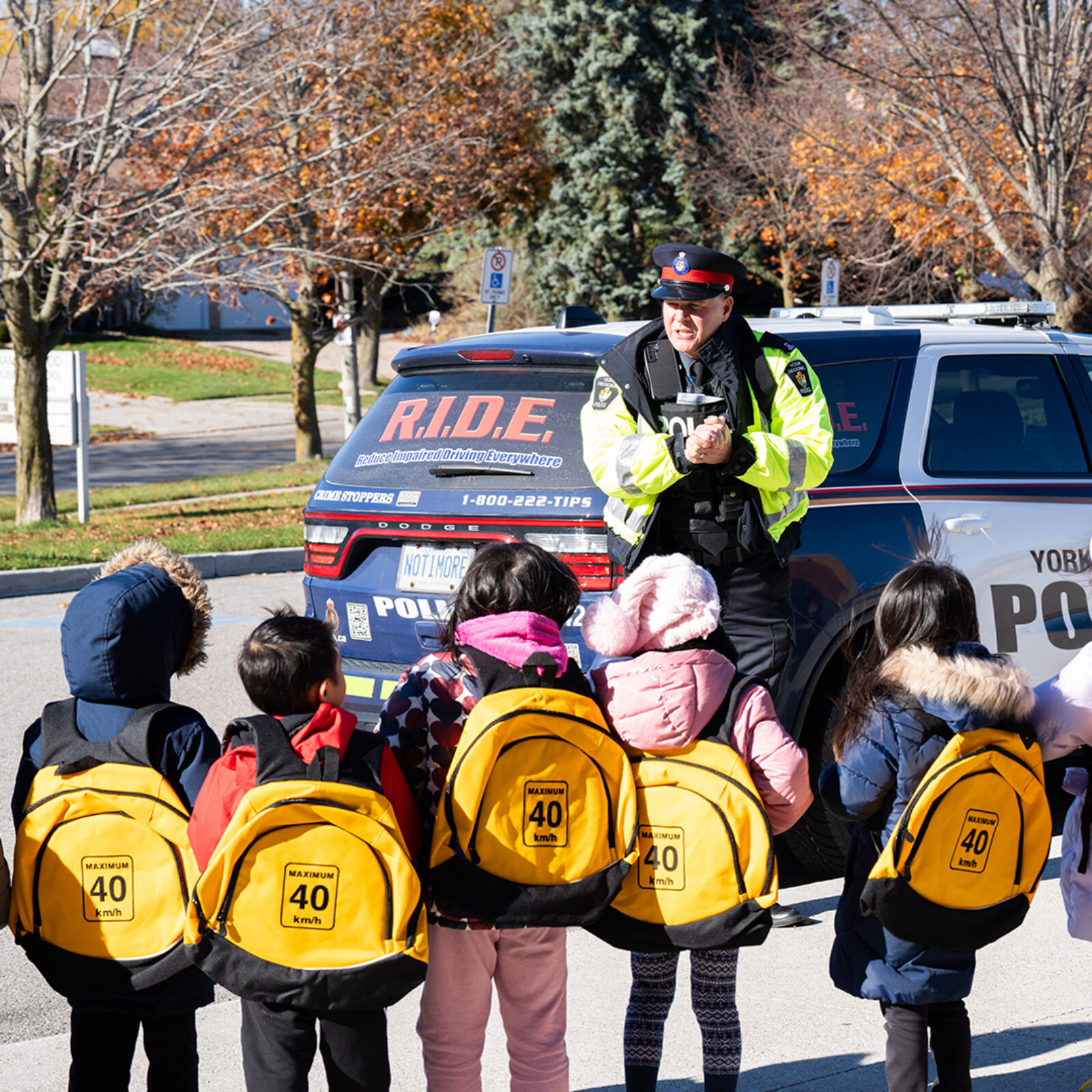 York Regional Police officer teaching children about road safety. The children are wearing yellow traffic backpacks labeled 'Maximum 40 km/h,' provided as giveaways by York Regional Police.