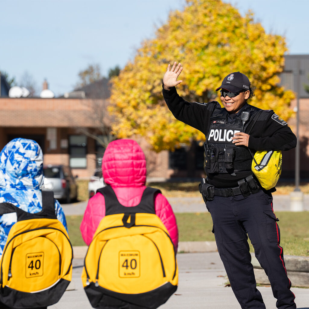 York Regional Police officer smiling and greeting children wearing bright yellow road safety backpacks labeled 'Maximum 40 km/h,' during a community safety education event.